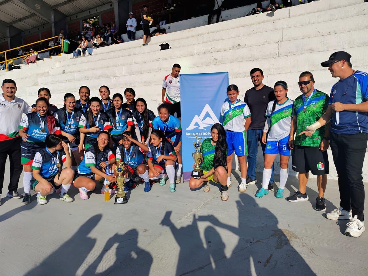 Equipo femenino de fútbol posando con trofeo y banner del Área Metropolitana en las graderías del estadio