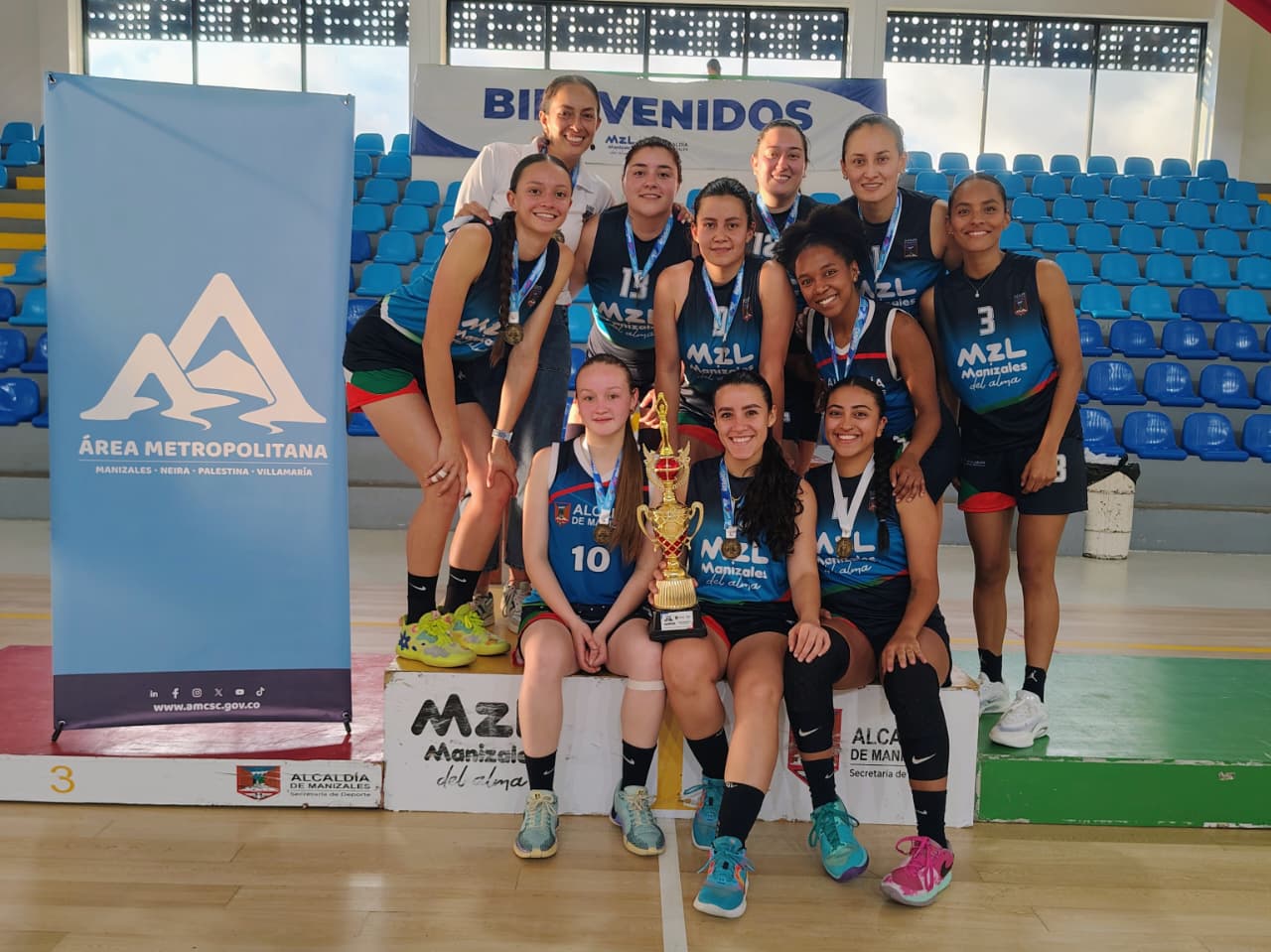 Equipo femenino de baloncesto con medallas y trofeo junto al banner del Área Metropolitana en coliseo cubierto